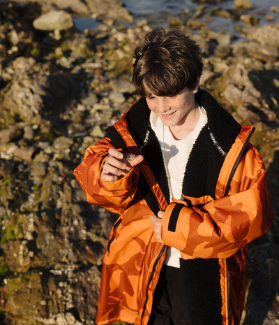 *MALE* A smiling child wearing a bright orange and brown camouflage hooded Dryrobe with a black zipper and the brand name 'dryrob' on the chest, standing outdoors near water and rocks.