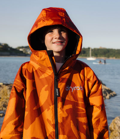 *MALE* A boy wearing a bright orange camouflage-patterned waterproof Dryrobe with black fleece lining, standing outdoors on a rocky shoreline.
