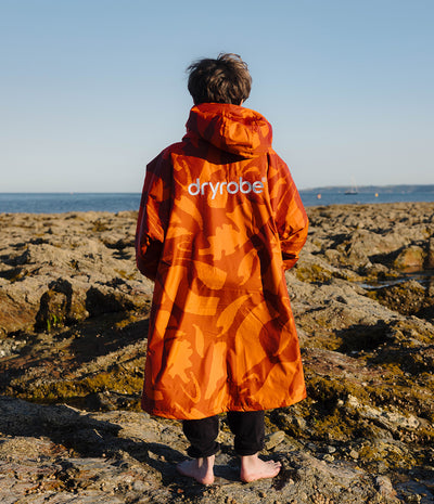 *MALE* A person standing on a rocky shoreline facing the ocean, wearing a bright orange hooded Dryrobe with a tonal abstract pattern and the brand name 'dryrobe' printed in white across the back.
