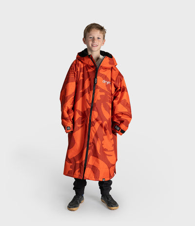 *MALE* A young boy modeling a bright orange and red camouflage hooded Dryrobe with black zippers and black cuffs, standing against a light gray studio background.