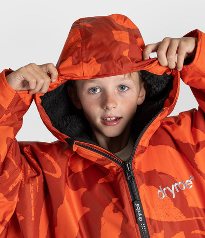*MALE* A child wearing an orange and red camouflage patterned hooded Dryrobe with a black fleece-lined hood, holding the hood up against a light gray studio background. The brand name 'dryroe' is visible on the chest and zipper pull.