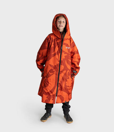 *MALE* A child model shown from the back wearing a vibrant orange and red camouflage hooded Dryrobe  with the brand name 'dryrobe' printed in white on the back, set against a plain light gray studio background.