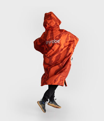 *MALE* A person wearing a bright orange and red camouflage hooded Dryrobe with the brand name 'dryrobe' printed in white on the back, paired with black pants and gray sneakers, captured mid-step against a light gray studio background.