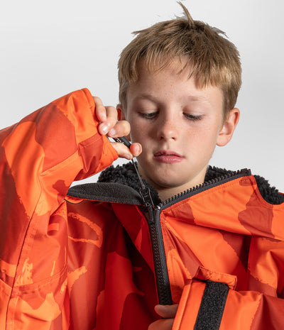 *MALE* A close-up of a young boy wearing a bright orange and red camouflage Dryrobe with black fleece lining and a black zipper, shown against a light gray studio background.