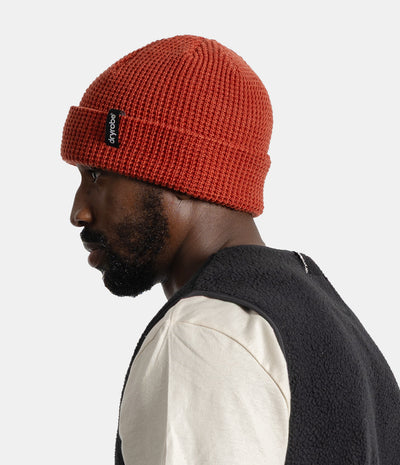 A man in profile wearing a textured orange-red knit beanie with a black logo patch on the cuff, paired with a cream shirt and a dark fleece vest, against a light gray studio background.