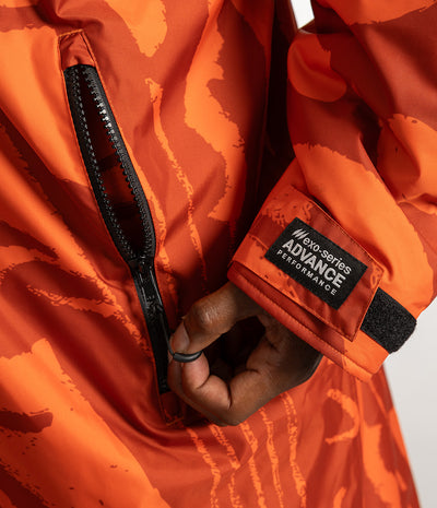 *MALE* Close-up of an orange patterned jacket with a black zipper and a visible sleeve patch reading 'no-series ADVANCE PERFORMANCE'. A hand is shown zipping the jacket.