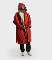 *MALE* A man wearing a hooded red changing robe with a white graphic print on the chest, layered over a white t-shirt, black pants, and beige sneakers, standing against a light gray studio background.