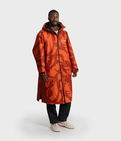 *MALE* A man modeling a long orange hooded parka with an abstract dark red-orange pattern, featuring a white 'dryrobe logo on the chest, black zippers, and black cuffs, paired with black pants and beige sneakers against a light gray background.