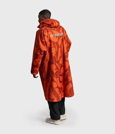*MALE* A model shown from the back wearing a long orange hooded raincoat with a tonal abstract pattern and the brand name 'dryrobe' printed in white on the upper back, paired with black pants and light-colored sneakers against a light gray studio background.