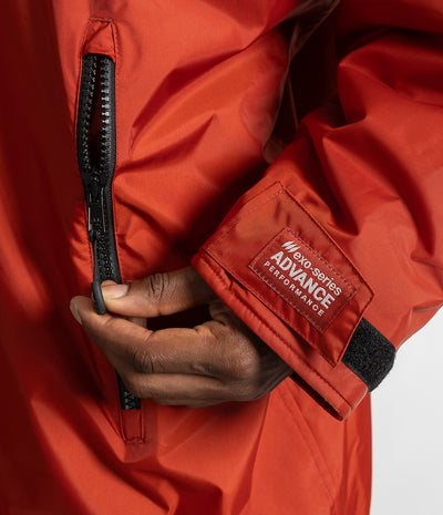 *MALE* Close-up of a person wearing a bright red technical Changing robe, pulling a black zipper on the chest while showing a branded cuff label that reads 'MAXI-SERIES ADVANCE PERFORMANCE'.