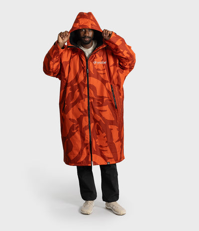 *MALE* A man wearing a long hooded raincoat with an orange and red abstract camouflage pattern, black pants, and beige sneakers, standing against a light gray studio background. The brand name 'dryrobe' is visible on the chest.