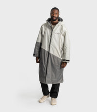 *MALE* A man wearing a long, color-blocked hooded raincoat in off-white and gray, with black pants and beige sneakers, standing against a light gray studio background.