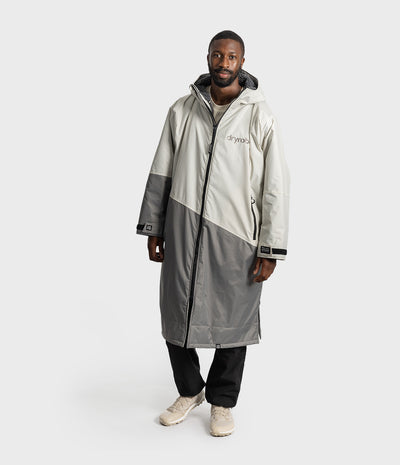 *MALE* A man wearing a long, color-blocked hooded raincoat in off-white and gray, with black pants and beige sneakers, standing against a light gray studio background.
