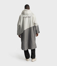 *MALE*A person shown from the back wearing a long hooded Dryrobe jacket in off-white and gray color blocking, paired with black pants and white sneakers, against a light gray studio background.