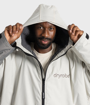 *MALE* A man wearing a light beige Dryrobot hooded jacket, holding the hood up with both hands against a clean white studio background.