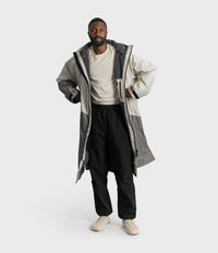 *MALE* A man modeling a long, oversized two-tone gray hooded jacket over a cream t-shirt, black pants, and beige sneakers, set against a plain light gray studio background.