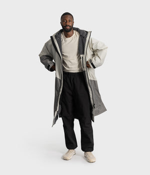 *MALE* A man modeling a long, oversized two-tone gray hooded jacket over a cream t-shirt, black pants, and beige sneakers, set against a plain light gray studio background.