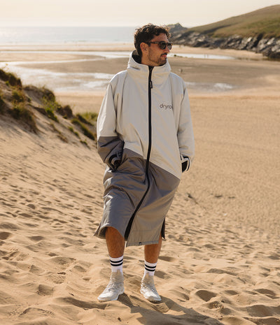*MALE* A man wearing a light gray and dark gray split-color hooded windbreaker jacket with a black zipper, standing on a sandy beach dune with the ocean and cliffs in the background.