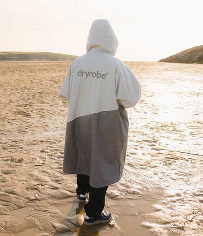 *MALE* A person standing on a sandy beach at sunset wearing a color-blocked hooded rain poncho with 'dryrobe' printed on the back. The poncho is white on the upper half and gray on the lower half.