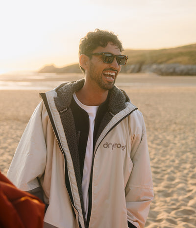 *MALE* A man wearing a beige Dryrobe waterproof jacket with a dark gray sherpa lining, black sunglasses, and a white t-shirt, standing on a sandy beach during golden hour.