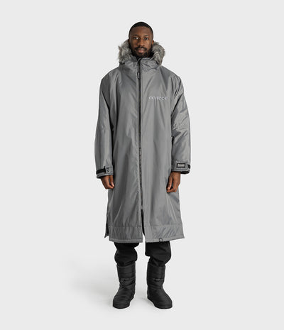 *MALE* A man wearing a long gray hooded Dryrobe with faux-fur trim, black pants, and black boots, standing against a light gray studio background. The parka has a visible 'dryrobe' logo on the left chest.