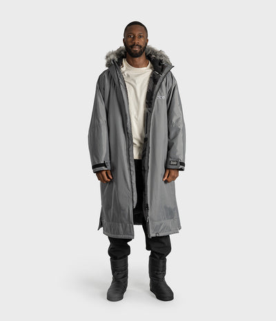 *MALE* A man modeling a long gray hooded Dryrobe with a faux-fur trimmed hood, worn over a cream sweatshirt, black pants, and black boots, against a light gray studio background.