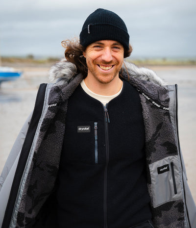 *MALE* A smiling man wearing a black beanie and a black zip-up fleece over a black Dryrobe with gray camouflage panels, gray faux-fur trim, and a white undershirt, standing outdoors on a beach with an overcast sky.