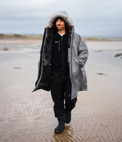*MALE* A man walking on a sandy beach wearing a long gray hooded Dryrobe with a black and gray patterned lining, a black zip-up hoodie, black pants, and black boots.