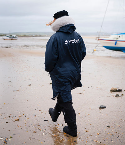 *MALE* A person walking on a muddy beach from behind, wearing a navy blue Dryrobe waterproof changing robe with a white faux-fur trimmed hood, black boots, and a black beanie. Boats and a cloudy sky are visible in the background.