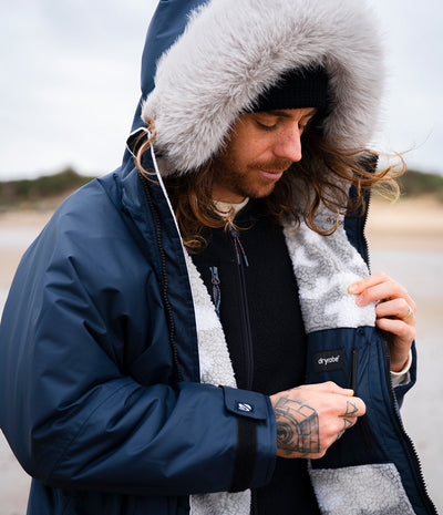 *MALE* A man wearing a navy blue Dryrobe with a fluffy gray faux-fur trimmed hood, a black beanie, and a black shirt, standing outdoors near a beach. He is opening the jacket pocket.