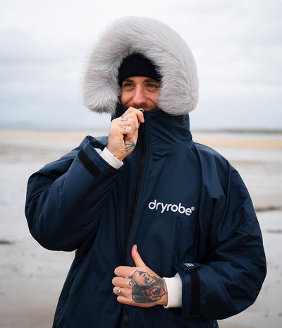 *MALE* A man wearing a navy blue Dryrobe with a large white faux-fur trimmed hood, a black beanie, and a black-and-white striped knit sweater, standing on a beach with his hands in his pockets.