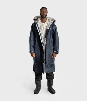 *MALE* A man modeling a long navy blue parka with a light gray faux-fur lined hood, worn over a white t-shirt, black pants, and black boots, against a light gray studio background.