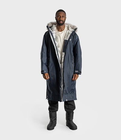 *MALE* A man modeling a long navy blue Dryrobe with a light gray faux-fur lined hood, worn over a white t-shirt, black pants, and black boots, against a light gray studio background.