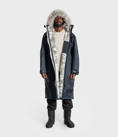 *MALE* A man modeling a long navy winter Dryrobe with a large gray faux-fur trimmed hood, worn over a cream shirt, black pants, and black boots, against a light gray studio background.
