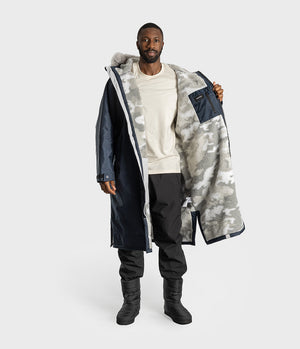 *MALE* A man modeling a long hooded parka with a navy blue exterior, white and gray camouflage interior lining, and a light gray background.
