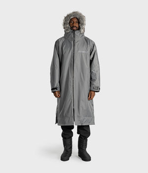 *MALE* A man wearing a long gray hooded parka with faux fur trim, black pants, and black boots, standing against a light gray studio background. The parka has a front zipper, zip pockets, and a small white logo on the left chest.