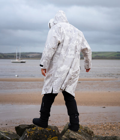 *MALE* A person standing on rocks at a beach, viewed from behind, wearing a hooded waterproof jacket in a light gray and white camouflage pattern, black pants, and black boots under an overcast sky.