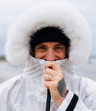 *MALE* A close-up portrait of a smiling man wearing a white polar-style Dryrobe with a large fluffy white fur-trimmed hood, a black knit beanie, and a white camouflage outer layer, with his zipper pulled up over his mouth.