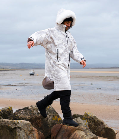 *MALE* A person wearing a white hooded waterproof Dryrobe, black pants, and black boots walks carefully across large rocks on a beach with shallow water and a cloudy sky in the background.