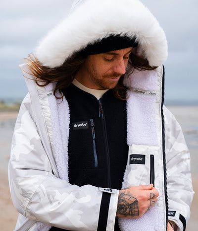 *MALE* A man wearing a white and gray camouflage Dryrobe with a white sherpa lining, a black fleece vest, and a white faux-fur lined hood, standing outdoors against a cloudy sky.