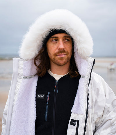 *MALE* A man wearing a white Dryrobe with a fluffy white fur-trimmed hood, a black fleece zip-up sweater, and a black beanie, standing outdoors against a cloudy, overcast sky.