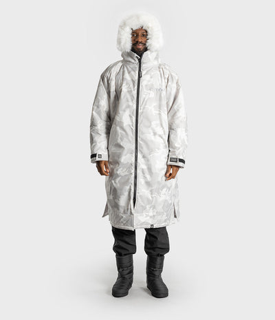 *MALE* A model wearing a long, hooded Dryrobe with a white camouflage pattern, a white faux-fur trimmed hood, black pants, and black boots, standing against a light gray studio background.