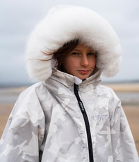 A person wearing a white and light gray camouflage hooded Dryrobe with a large fluffy white fur-trimmed hood, photographed outdoors against a muted overcast sky and distant landscape.