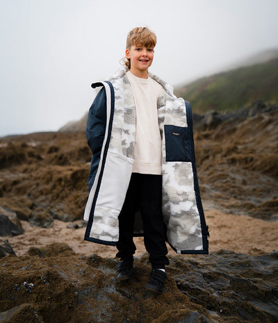 *MALE* A child stands on a rocky beach wearing a long navy and white camouflage-patterned Dryrobe over a cream sweatshirt and black pants, with misty hills in the background.