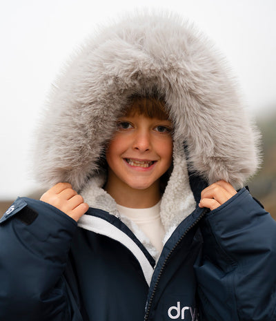 *MALE* A smiling young boy wearing a navy blue winter Dryrobe with a large fluffy light gray faux-fur hood trim and a white fleece lining, holding the hood open against an overcast sky.