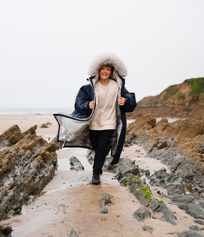 *MALE* A child walking on a rocky beach path wearing a navy blue hooded Dryrobe with a large white faux-fur trim, a cream sweatshirt, and black pants.