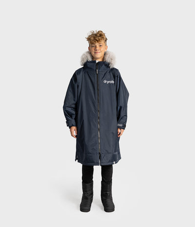 *MALE* A child model wearing a navy blue Dryrobe kids' with a light gray faux-fur hood trim, black pants, and black boots, standing against a light gray studio background.
