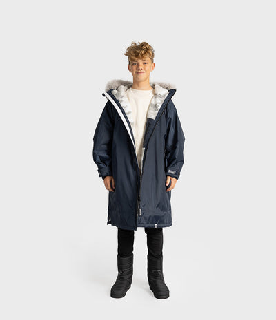 *MALE* A child model wearing a navy blue hooded parka with a light gray and white faux fur trim, a cream shirt, black pants, and black boots, standing against a light gray studio background.
