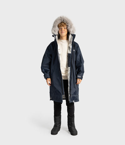 *MALE* A child model wearing a navy blue hooded Dryrobe with a light gray faux-fur trim, layered over a cream sweater, black pants, and black boots, set against a plain light gray studio background.