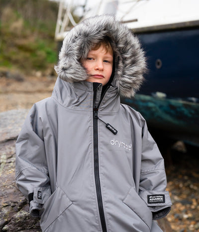 *MALE* A child wearing a gray Dryrobe waterproof jacket with a faux-fur trimmed hood, standing outdoors in front of a boat.